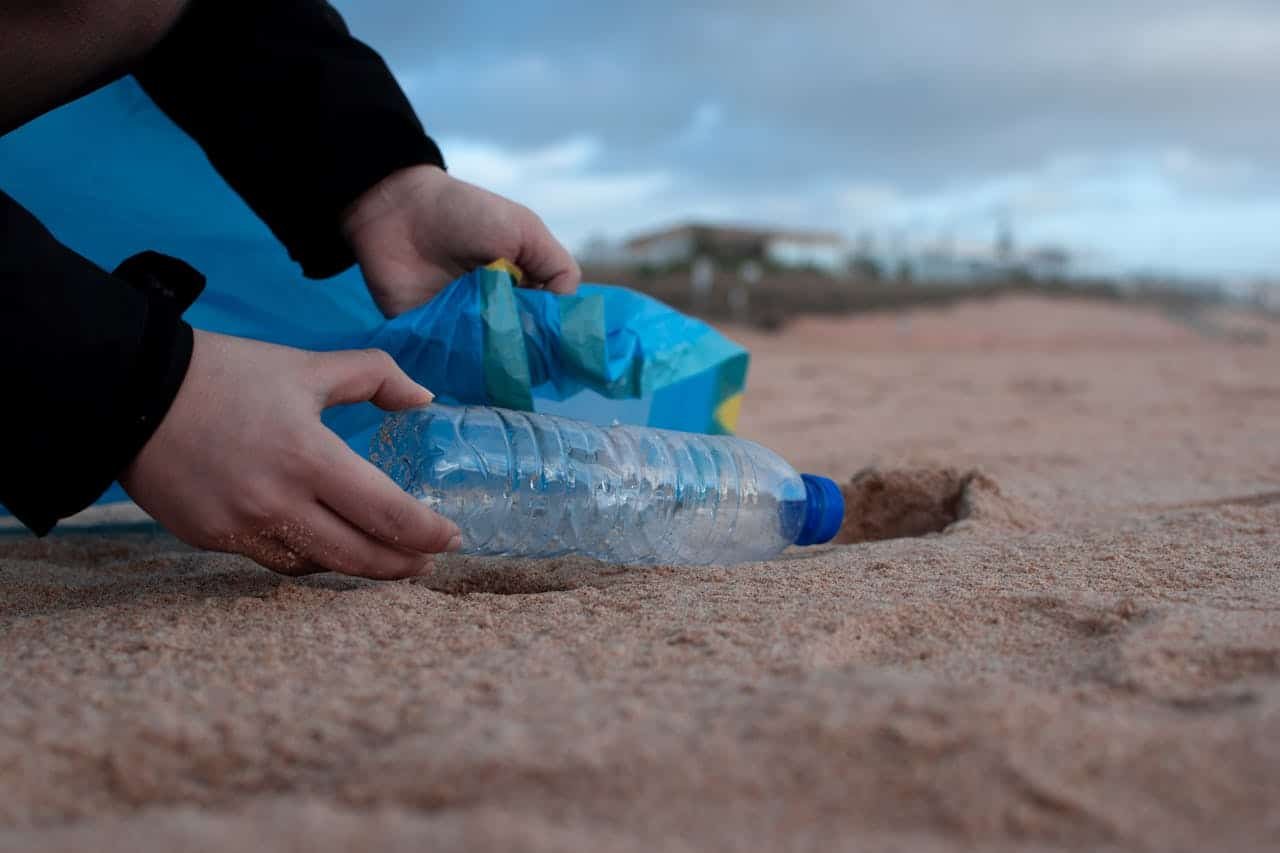 hero-img-01 Hands collecting a plastic water bottle from the sandy beach, promoting environmental awareness.